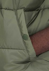 Olivgrüne isolierte Steppjacke mit einer genähten Tasche, ausgestattet mit einem dunkelgrünen Druckknopf und einer glatten, glänzenden Textur.