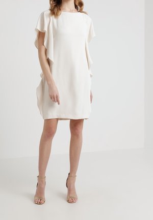 Femme vêtue d'une robe blanche courte, à manches courtes, avec des détails à volants et des talons beiges ouverts à la cheville, debout devant un fond blanc uni.