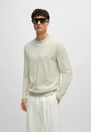 Pullover in maglia beige chiaro con collo e orlo a coste, caratterizzato da un logo discreto sul petto. Il modello indossa occhiali da sole scuri e pantaloni chiari.