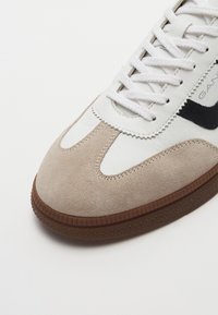 Sneaker featuring white leather and beige suede upper, black accent, round toe, and textured brown rubber sole with star pattern.