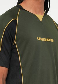 Tamsiai žalia sportinė marškinėliai su juodomis rankovėmis, geltonais akcentais ir "UMBRO" logotipu. Textūruotas audinys su V formos iškirpte.