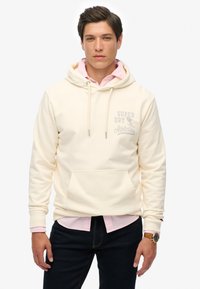 Crème hoodie met een trekkoord, voorzak en grijs "Super Dry Athletics" logo. Draagt over een roze shirt met kraag en donkere jeans.