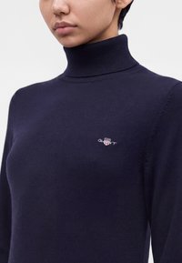 Femme portant un pull col roulé bleu marine cintré avec un petit logo GANT brodé sur la poitrine.