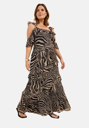 Vrouw draagt een vloeiende zebra-print jurk met geplooide lagen, cold-shoulder mouwen en zwarte sandalen met open neus, staand tegen een witte achtergrond.