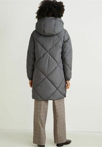 Grå pufferjakke med hætte, der har diagonale quiltede mønstre og elastiske manchetter, sammen med beige ternede bukser og mørke sko.
