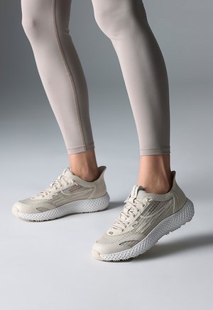 Person trägt beige Leggings und hellbeige Sportschuhe mit strukturierten weißen Sohlen, steht vor einem neutralen grauen Hintergrund.