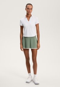 Biała koszulka polo z dekoltem w kształcie litery V, w zestawieniu z krótką, zieloną plisowaną spódnicą z białym obszyciem. Model nosi białe buty sportowe i skarpetki.