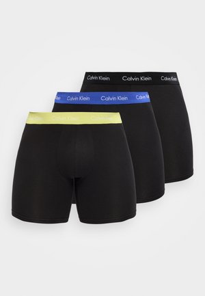 Drei schwarze Boxer-Slips in einer Reihe mit Calvin Klein gebrandeten Taillenbändern in Schwarz, Blau und Gelb von vorne nach hinten.