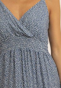 Robe bleue avec un décolleté en V, smocké froncé à la taille, et un tissu doux présentant un motif de feuilles blanches sur l'ensemble.