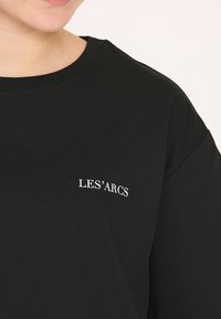 Sort bomulds T-shirt med rund halsudskæring og korte ærmer. Har hvid tekst "LES'ARCS" broderet på brystet. Glat tekstur.