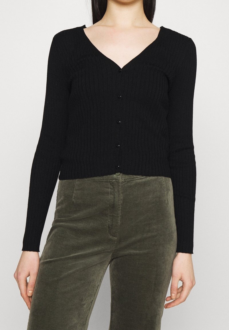 Svart ribbad cardigan med V-ringning, långa ärmar och knäppning fram. Bärs över olivgröna kordbyxor som visar en strukturerad design.