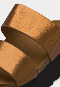 Κοντινό πλάνο ενός χρυσού μεταλλικού σανδαλιού με διπλό λουράκι, πάτο από φελλό και μαύρη σόλα πλατφόρμας.