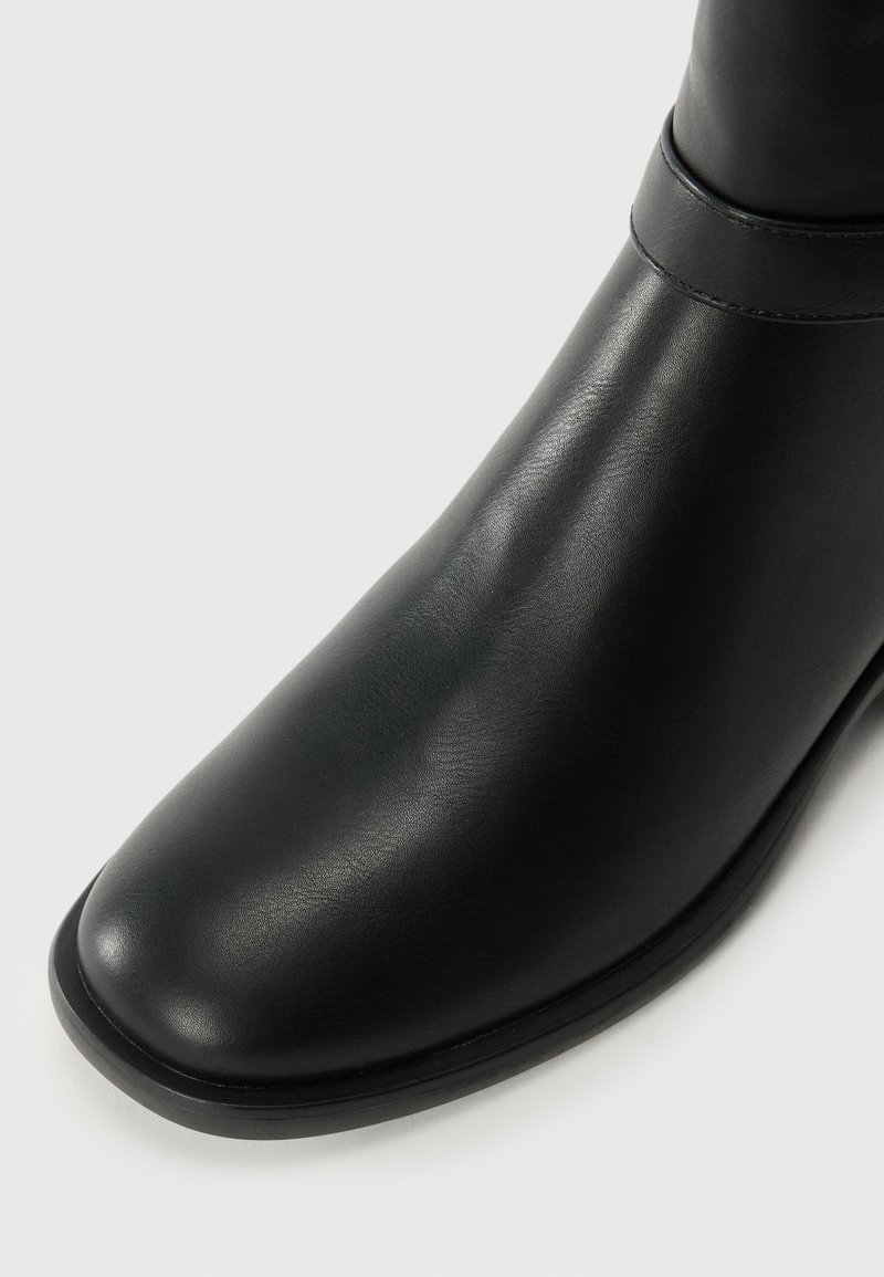 Bota de cuero negro hasta el tobillo con una textura suave, punta redondeada y costuras mínimas. Presenta un ligero tacón y un diseño elegante.