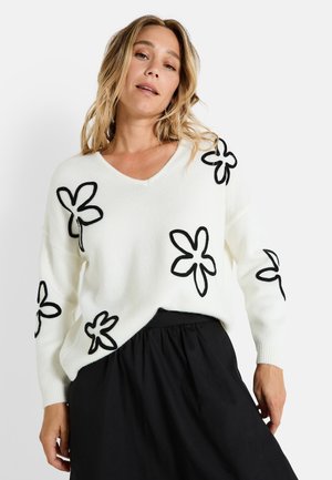 Pull en tricot blanc à col en V, orné d'appliqués floraux noirs. Associé à une jupe noire, mettant en avant une coupe décontractée et une texture.