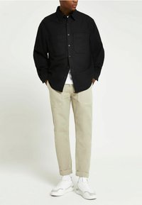 Czarna, długa koszula zapinana na guziki z dwoma kieszeniami, w połączeniu z jasnobeżowymi spodniami i białymi trampkami z szarymi akcentami.