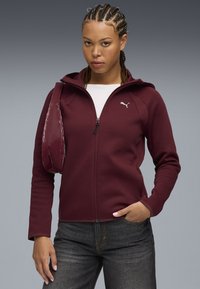 Burgundy zip-up hoodie made from soft fabric with a fitted design. Features a white logo on the chest and a pocket on the sleeve.