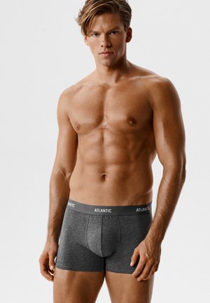 Męski, muskularny mężczyzna z krótkimi włosami, ubrany w ciemnoszare bokserki Atlantic, stoi na jasnym, jednolitym tle.