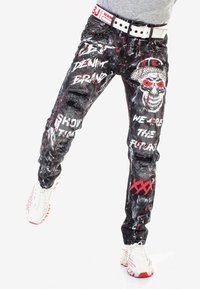 Abgenutzte schwarze Jeans mit rotem und weißen Spritzdesign, versehen mit einem Totenkopf-Motiv und auffälligem Text. Enthält einen weißen Gürtel mit roten Akzenten.