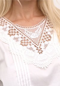 Bílý top s jemným krajkovým zdobením v oblasti výstřihu, s květinovými vzory a geometrickými motivy; má plisované přední části.