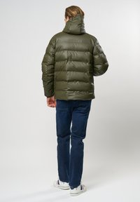 Alyvuogių žalia pūkų striukė su aukštu apykakle, pasižyminti blizgiu paviršiumi ir tolygiai išdėstytu horizontaliai suknežintu raštu. Užsidėta ant mėlynų džinsų ir sportinių batelių.