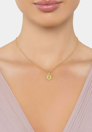 Nő aranylánc nyakláncot visel, amelyen egy négyzet alakú sárga drágakő medál található, kisebb áttetsző kövekkel körülvéve, világos felsőruházat ellen.