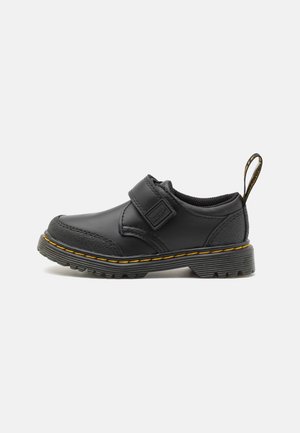 Zapato negro para niño pequeño con tira de velcro, costuras amarillas a lo largo de la suela, puntera texturizada y lengüeta en el talón sobre un fondo blanco.