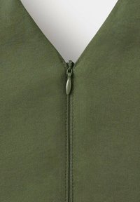 Gros plan d'un vêtement en tissu vert avec une fermeture éclair centrée et fermée et un col en V sur fond blanc.