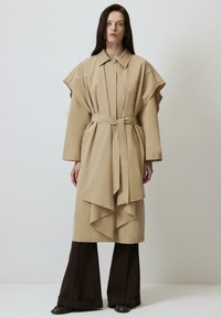Palton bej cu curea, mâneci supradimensionate și tiv în straturi. Asortat cu pantaloni largi negri și botine, design minimalist.