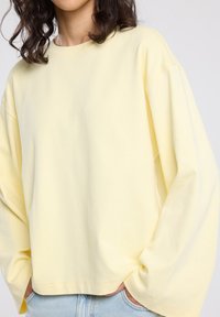 Person wearing a pale yellow oversized long-sleeve shirt with hands in light blue denim jeans pockets, showing upper torso and curly hair.