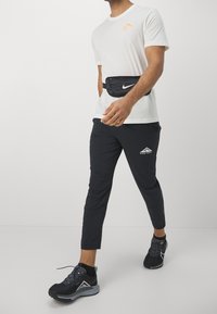 Camisa blanca de manga corta con logo naranja, riñonera negra con cremallera, pantalones negros ajustados con logo, y zapatillas deportivas negras con detalles grises.