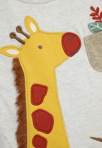 Design de girafe jaune avec des accents marron et une crinière texturée sur un fond en tissu gris clair ; présente des taches rouges et des détails faciaux simples.