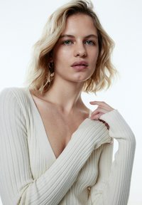 Helles beige, geripptes Sweater mit tiefem V-Ausschnitt, langen Ärmeln und weicher Textur. Teilweise sichtbares Schlüsselbein und glatte Haut.