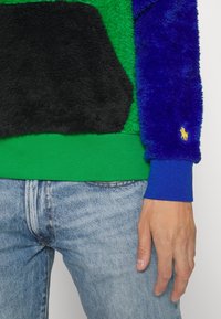 Fleecová mikina s černými a zelenými panely, modrými rukávy, žebrovaným zeleným lemem a malým žlutým logem. Jemná struktura po celém povrchu.