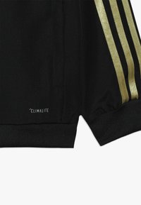 Black fabric with three gold stripes and climacool logo near the hem, featuring ribbed cuffs and a small side slit.