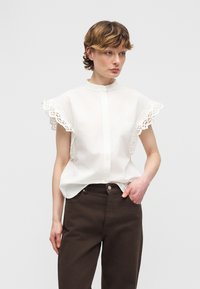 Femme aux cheveux courts portant un chemisier blanc avec des manches à volants en dentelle et un pantalon taille haute marron foncé, debout avec une main dans la poche.