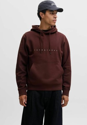 Jeune homme portant un sweat à capuche marron foncé JACK & JONES et une casquette noire, regardant sur le côté sur un fond clair uni.