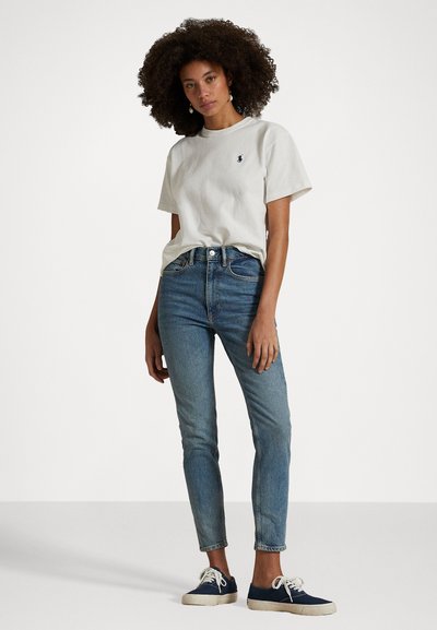 Mulher com cabelo cacheado, vestindo uma camisa branca de manga curta, jeans de cintura alta azul e ténis azul-marinho, de pé contra um fundo simples.