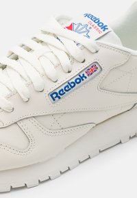 Baltas Reebok Classic sportbačių modelis su lygiu odiniu paviršiumi, apvaliu nosiu ir tekstūruota guma. Funkcijos: mėlynas logotipas ir anglijos vėliavos akcentas.