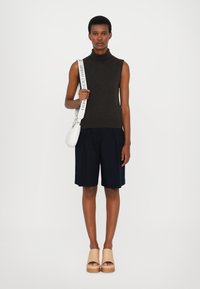 Mørkebrun ermeløs høyhalset genser, marineblå plissert shorts, beige klosser. I tillegg en hvit skulderveske med logobånd holdt i hånden.