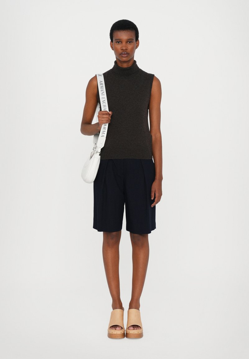 Mørkebrun ermeløs høyhalset genser, marineblå plissert shorts, beige klosser. I tillegg en hvit skulderveske med logobånd holdt i hånden.