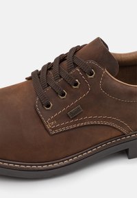 Scarpa in pelle marrone con una finitura liscia, forma arrotondata, cinque asole e lacci marroni scuri. Presenta cuciture a contrasto e un accento con marchio.