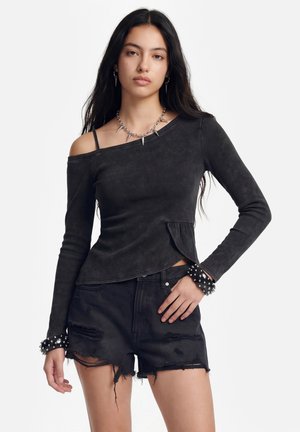 Nuori nainen, jolla on pitkät tummat hiukset ja joka pukeutuu epäsymmetriseen mustaan pitkähihaiseen toppiin, revittyihin mustiin shortseihin, piikki-rannekoruihin ja piikkikaulakoruun.