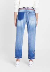 Jeans de mezclilla azul claro con un ajuste holgado, que cuentan con un parche azul oscuro en la parte trasera, dobladillos deshilachados y texto bordado cerca del puño.