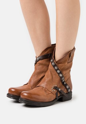 Botas de cuero marrón hasta el tobillo con acentos de tachuelas metálicas, un diseño plegado y un tacón bajo negro. Presentan una correa lateral y un detalle de marca.