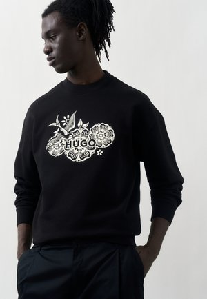 Fiatal férfi dreadlockkal, fekete pulóvert visel fehér virágmintákkal és a 