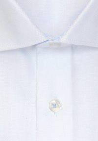 Gros plan sur le col et la patte de boutonnage avant d'une chemise blanche, présentant un bouton surpiqué en haut et un second bouton transparent en dessous.