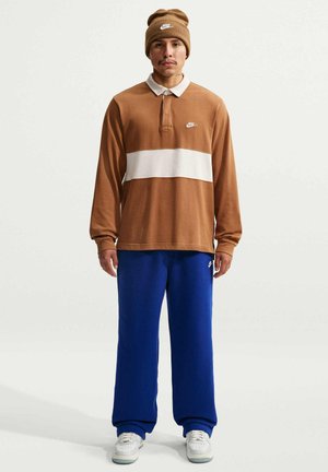 Mężczyzna ubrany w brązowo-białą długą koszulkę polo Nike, niebieskie spodnie dresowe Nike, białe trampki i brązową czapkę Nike, stojący na jednolitym tle.