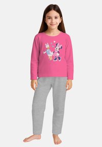 Ragazza sorridente che indossa una maglietta a maniche lunghe rosa con stampa di Minnie e Paperina e pantaloni della tuta grigio chiaro, in piedi a piedi nudi su uno sfondo bianco.