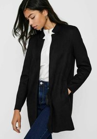 Giacca lunga in suede nera con colletto, tasche laterali e vestibilità dritta, abbinata a una blouse bianca e jeans scuri.
