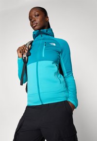 Giacca in pile color teal con colletto alto e tasche con zip, caratterizzata da un pannello di teal scuro in contrasto. Indossata con pantaloni cargo neri e uno zaino.